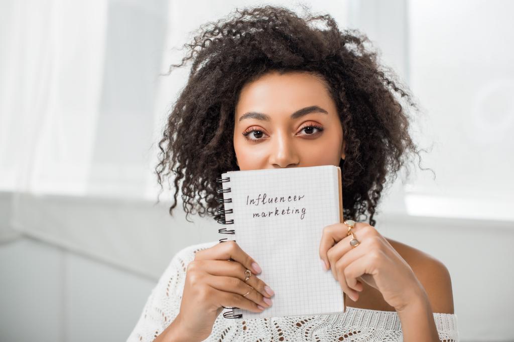 кучерява афроамериканська дівчина тримає блокнот з маркетинговим написанням впливу вдома
  - Фото, зображення