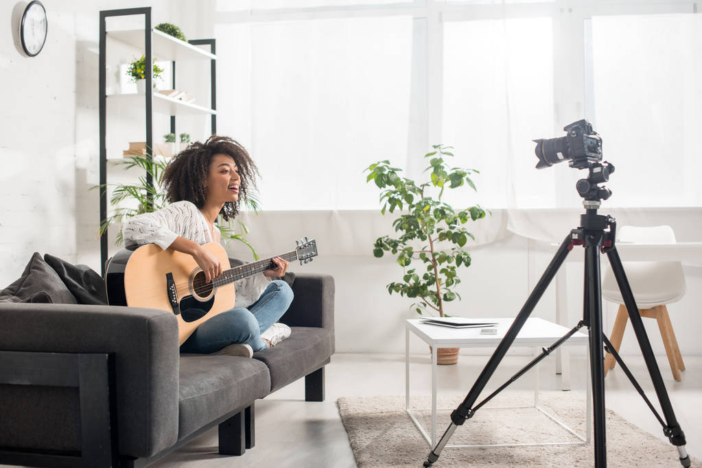 felice ragazza afro-americana in bretelle suonare la chitarra acustica vicino alla fotocamera digitale
  - Foto, immagini