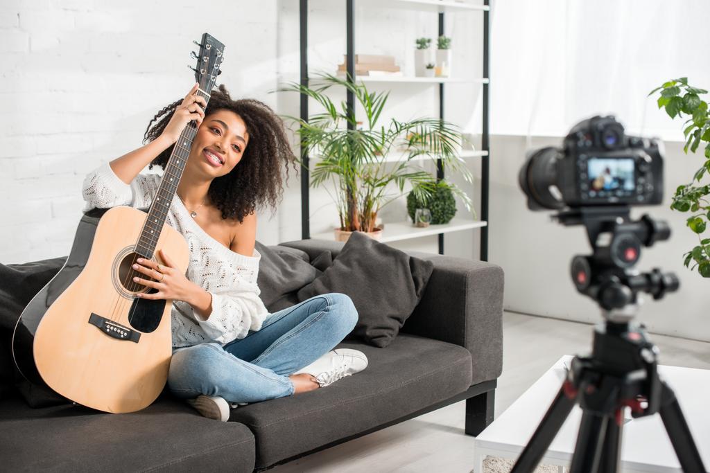 επιλεκτική εστίαση του χαρούμενου Αφροαμερικανού κοριτσιού σε τιράντες κρατώντας ακουστική κιθάρα και κοιτάζοντας ψηφιακή φωτογραφική μηχανή  - Φωτογραφία, εικόνα