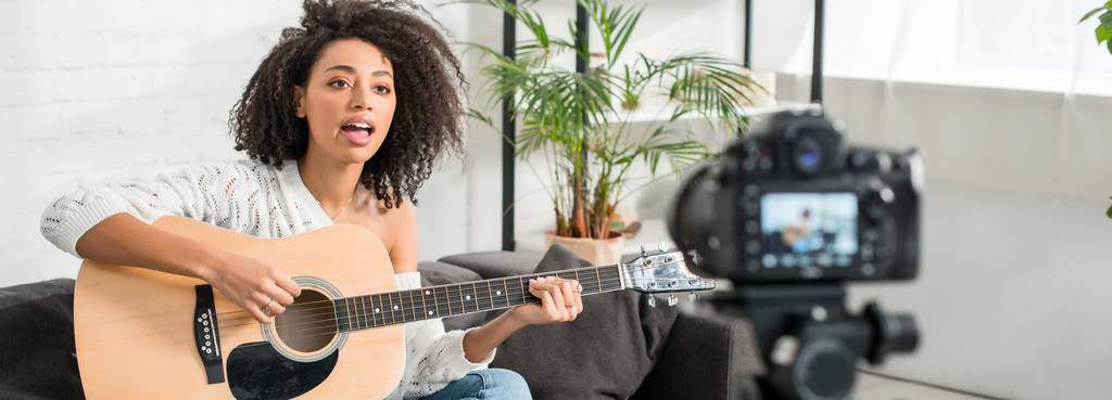 scatto panoramico di giovane ragazza afro-americana in bretelle suonare la chitarra acustica e cantare vicino alla fotocamera digitale
  - Foto, immagini