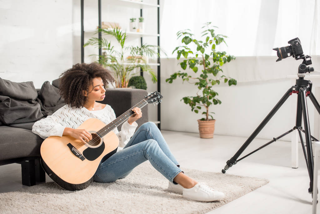 foco seletivo de jovem e encaracolado menina americana africana tocando guitarra acústica perto de câmera digital na sala de estar
  - Foto, Imagem