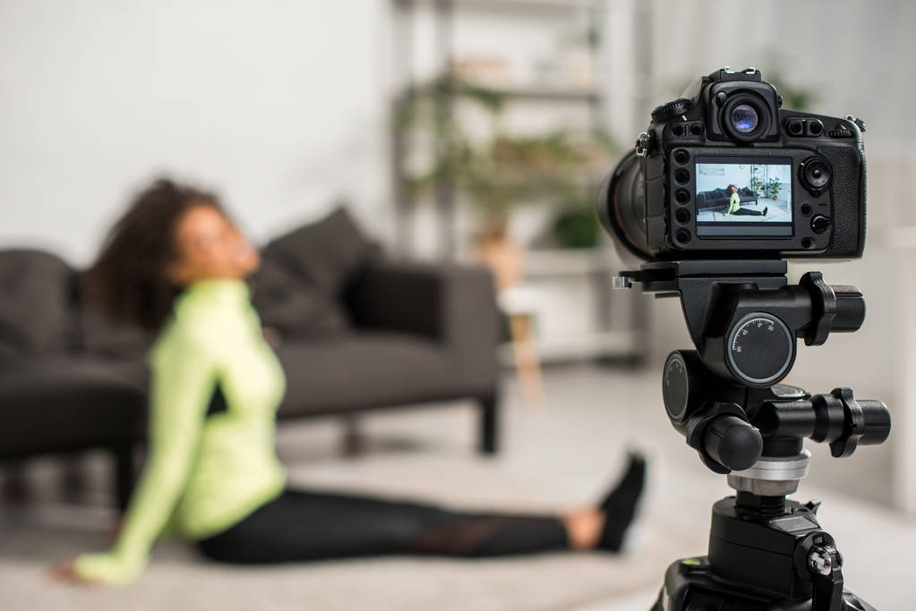 επιλεκτική εστίαση της ψηφιακής φωτογραφικής μηχανής με κουρασμένο αφροαμερικανό blogger βίντεο σε τιράντες ακουμπισμένη στο χαλί στην οθόνη  - Φωτογραφία, εικόνα