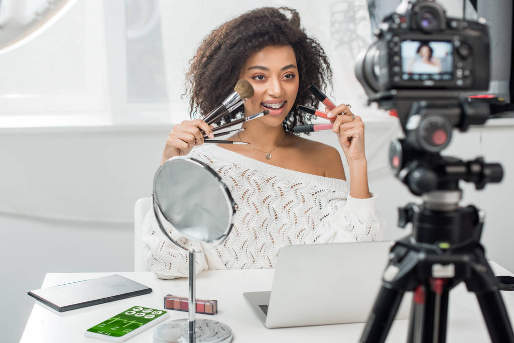selektiver Fokus des glücklichen afrikanisch-amerikanischen Influencers in Zahnspangen, die Lippenglanz und Kosmetikbürsten in der Nähe des Smartphones mit Fitness-Tracker und Digitalkamera halten - Foto, Bild