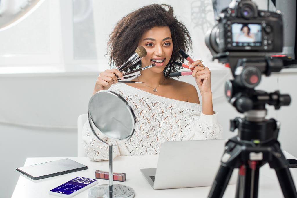 selective focus of happy african american influencer in braces holding lip glosses and cosmetic brushes near smartphone with medical tracker and digital camera - Photo, Image