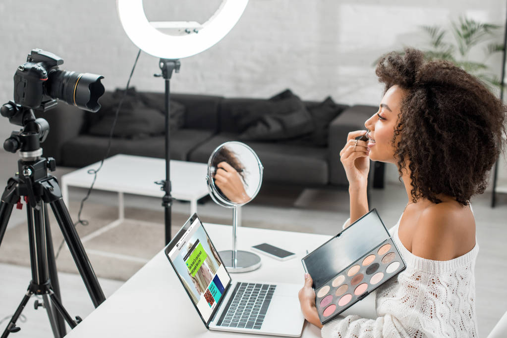 KYIV, UKRAINE - DECEMBER 10, 2019: side view of african american influencer holding eye shadow palette near laptop with bbc website and digital camera  - Photo, Image
