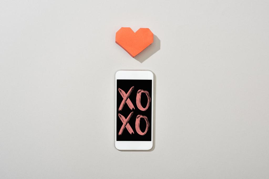 Bovenaanzicht van smartphone met xoxo illustratie en papieren hart op grijze achtergrond - Foto, afbeelding
