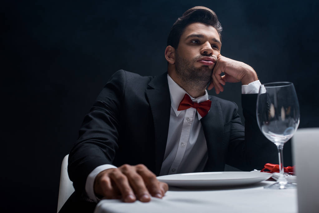 Selektivní zaměření pohledného muže v obleku sedí u servírovaného stolu izolovaný na černém - Fotografie, Obrázek