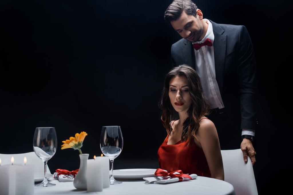 Όμορφος άντρας στέκεται πίσω από κομψή γυναίκα στο τραπέζι που σερβίρεται απομονωμένος στο μαύρο - Φωτογραφία, εικόνα