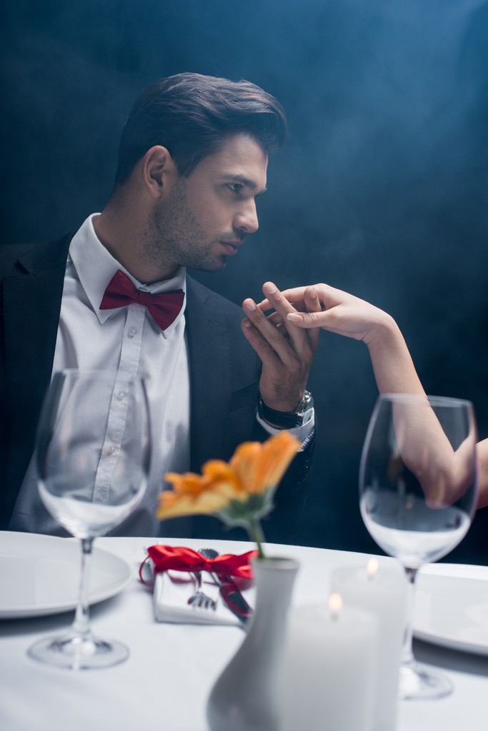 Vue latérale du bel homme tenant la main de la femme et regardant loin de la table servie sur fond noir avec de la fumée
 - Photo, image
