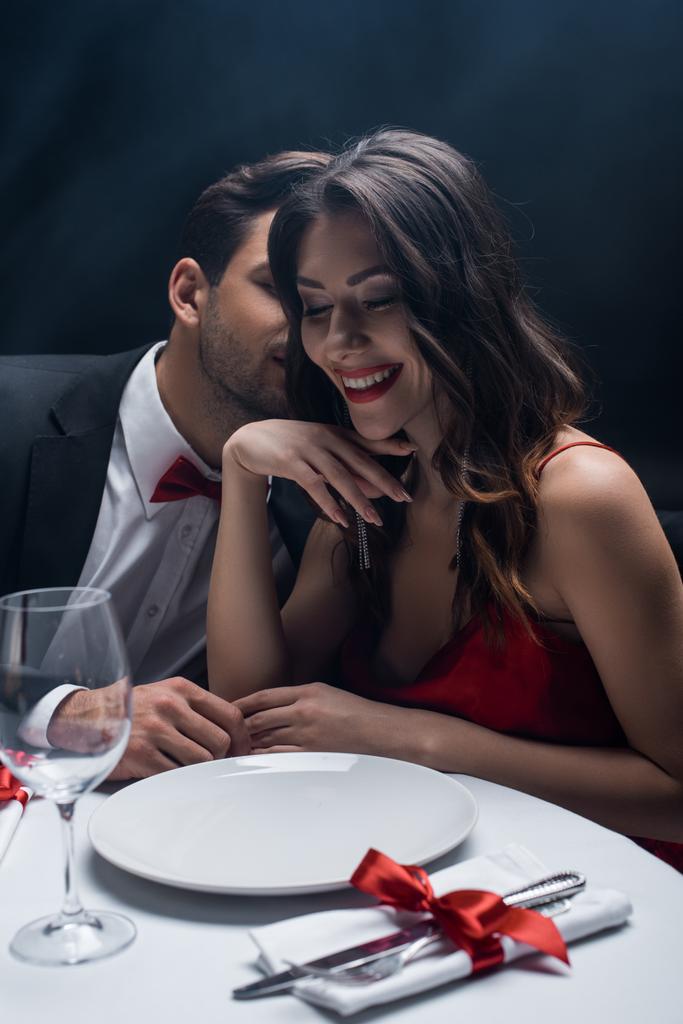 schöner Mann flüstert der schönen lächelnden Frau am Serviertisch auf schwarzem Hintergrund mit Rauch zu - Foto, Bild