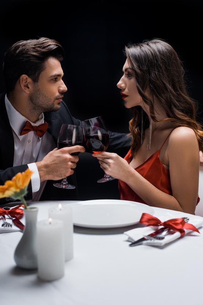 Vista lateral do casal elegante olhando um para o outro enquanto clica com vinho na mesa servida isolado em preto
 - Foto, Imagem