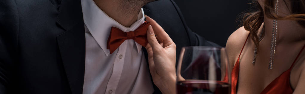 Обрізаний вид жінки, що регулює краватку чоловіка келихом вина ізольовано на чорному, панорамний знімок
 - Фото, зображення