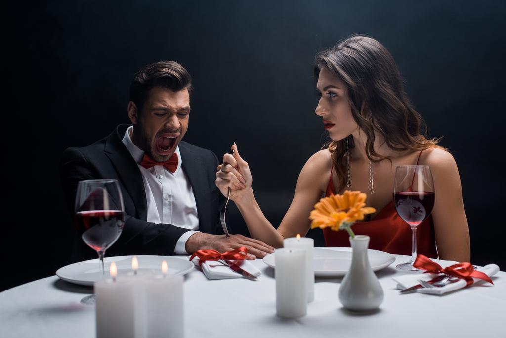 Όμορφη γυναίκα καρφώνει πιρούνι στο χέρι του άντρα ουρλιάζοντας κατά τη διάρκεια ρομαντικό δείπνο απομονωμένο σε μαύρο - Φωτογραφία, εικόνα