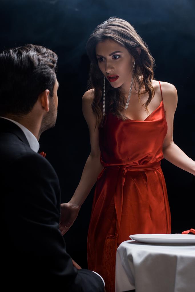 Wütende Frau blickt Freund am Serviertisch auf schwarzem Hintergrund mit Rauch an - Foto, Bild