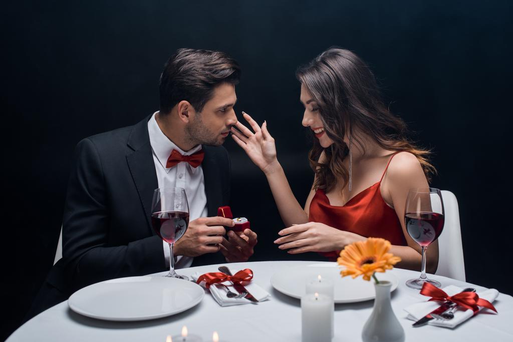 Seitenansicht des hübschen Mannes präsentiert Schmuckring an lächelnde Freundin beim romantischen Abendessen isoliert auf schwarz - Foto, Bild