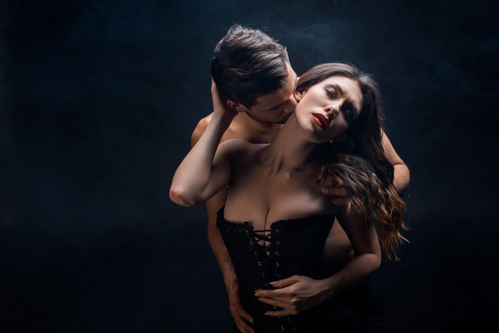 Shirtless man kissing in neck sexy woman in corset on black background with smoke - Photo, Image