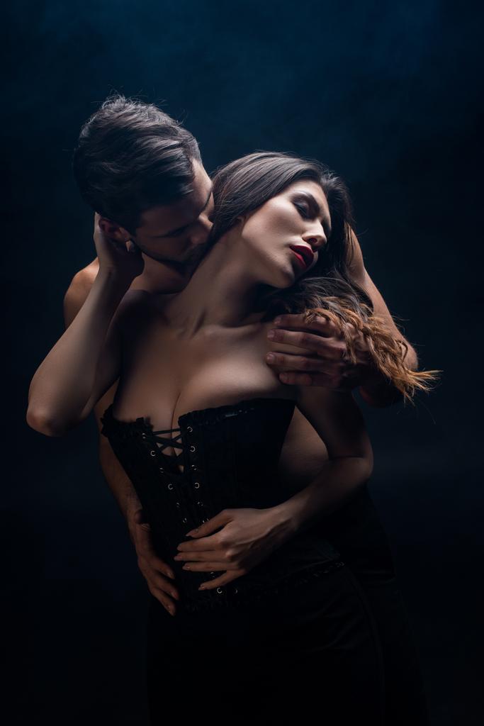 Hombre guapo sin camisa besándose en el cuello mujer atractiva en corsé aislado en negro
 - Foto, Imagen