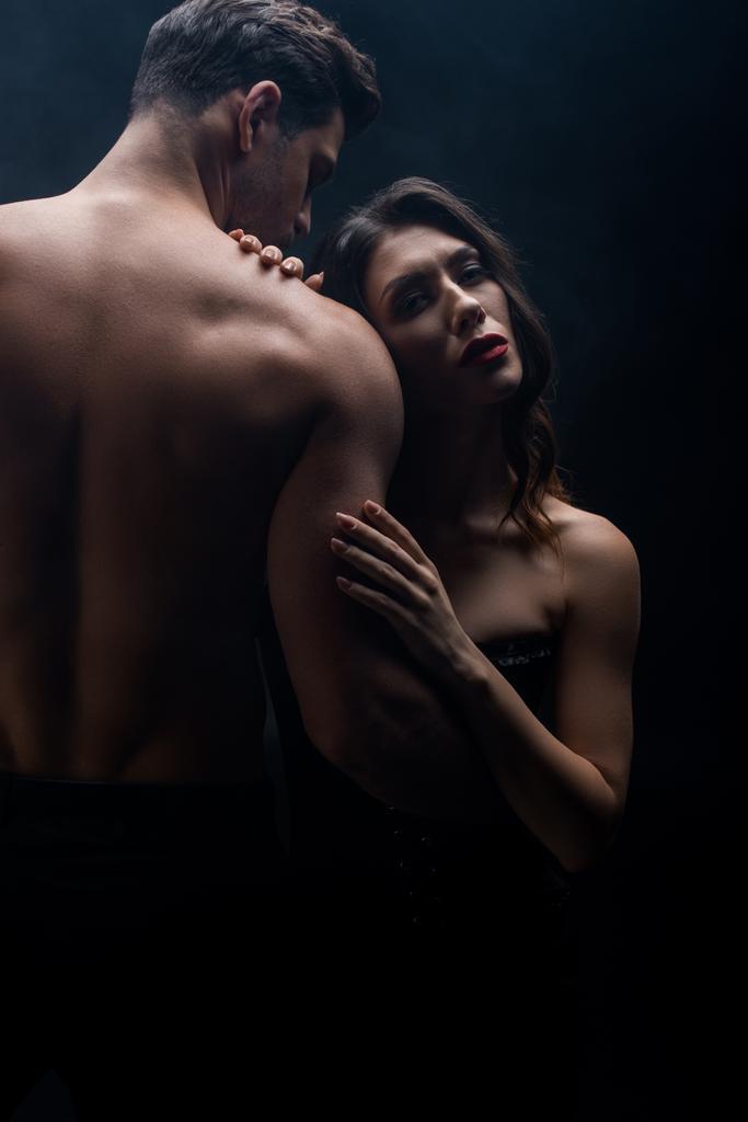Mujer atractiva abrazando novio sin camisa y mirando a la cámara aislada en negro
 - Foto, imagen