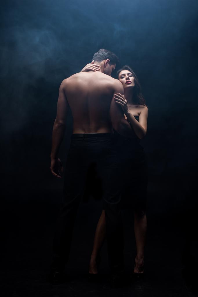 Повна довжина красивої жінки обіймає без сорочки чоловіка на чорному тлі з димом
 - Фото, зображення