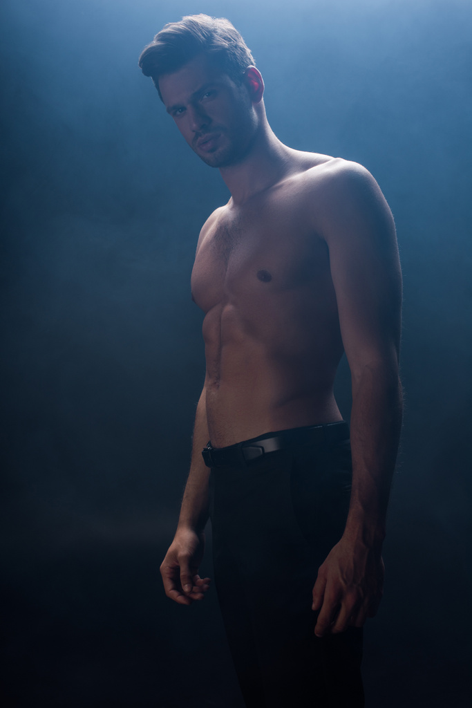 Низкий угол обзора красивого мускулистого человека, смотрящего на камеру на черном фоне с дымом
  - Фото, изображение