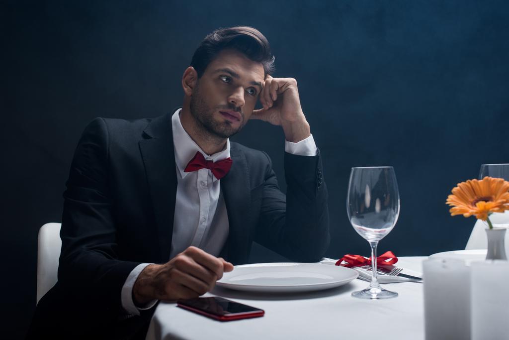 Елегантний чоловік з рукою по голові, сидячи за допомогою смартфона на поданому столі на чорному тлі з димом
  - Фото, зображення