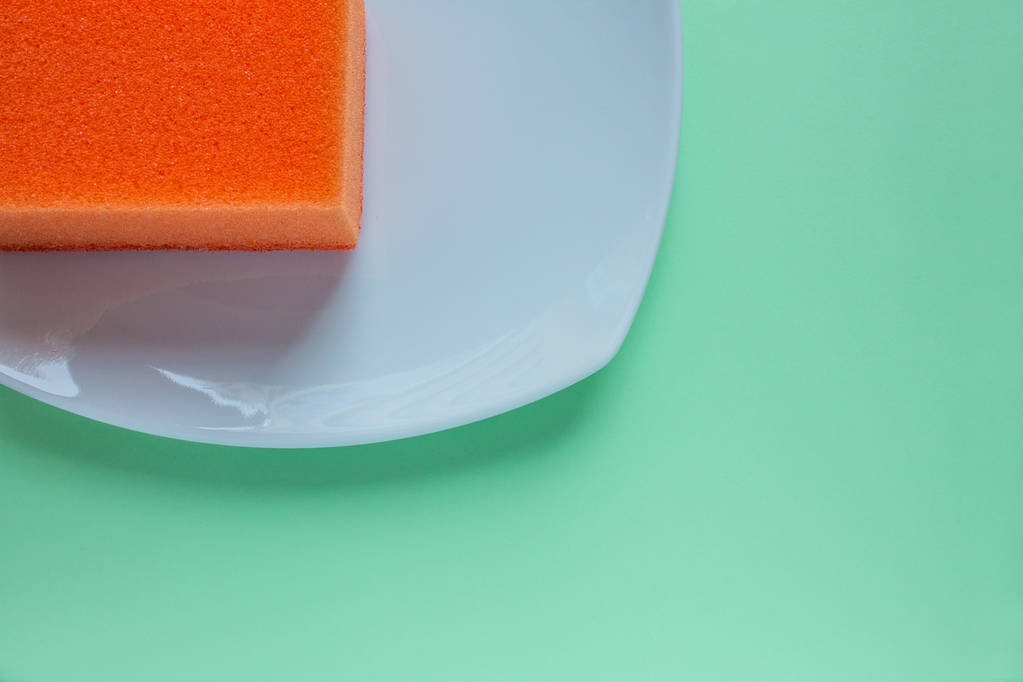 Pomarańczowa gąbka do zmywania naczyń na kwadratowym białym talerzu z bliska na równym tle miętowej zieleni. Koncepcja: czyste naczynia - Zdjęcie, obraz