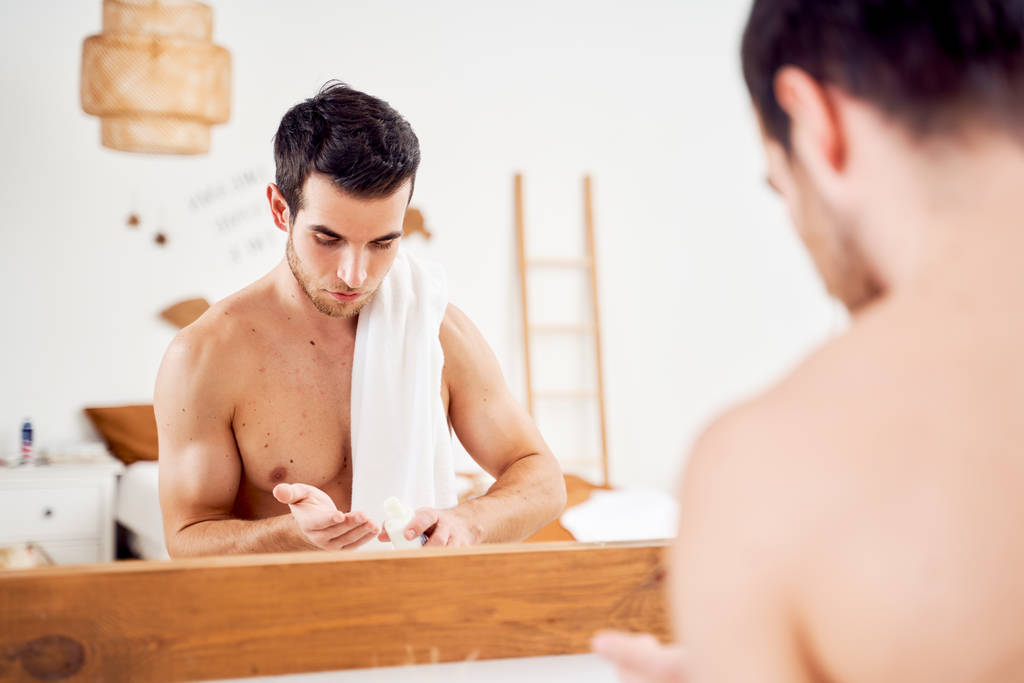 Young sports man with lotion in hands and towel on shoulders stands in front of bathroom mirror - Photo, Image