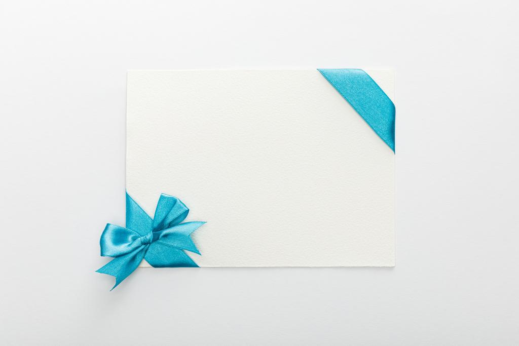青い装飾サテンリボンと白い背景の弓を持つ空白のカードのトップビュー - 写真・画像