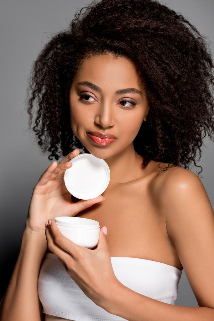 schöne afrikanisch-amerikanische Mädchen hält Plastikbehälter mit kosmetischer Creme, isoliert auf grau - Foto, Bild