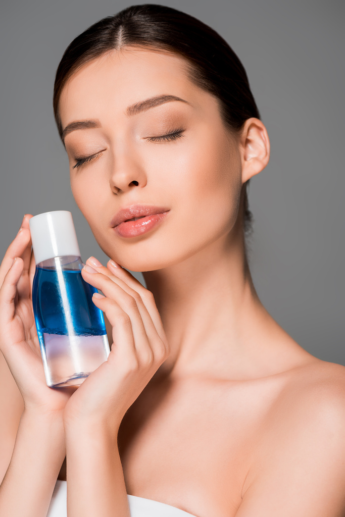 portrait of tender woman with closed eyes holding bottle of makeup remover, isolated on grey - Photo, Image