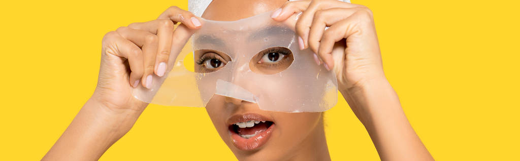 panoramic shot of surprised african american girl with towel on head applying hydrogel mask, isolated on yellow - Photo, Image