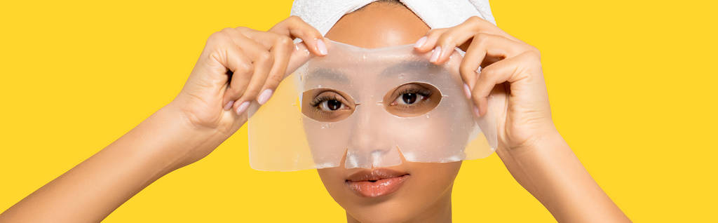 Kafasında havluyla sarıya izole edilmiş hidrojel maske takan çekici Afrikalı Amerikalı kızın panoramik çekimi. - Fotoğraf, Görsel