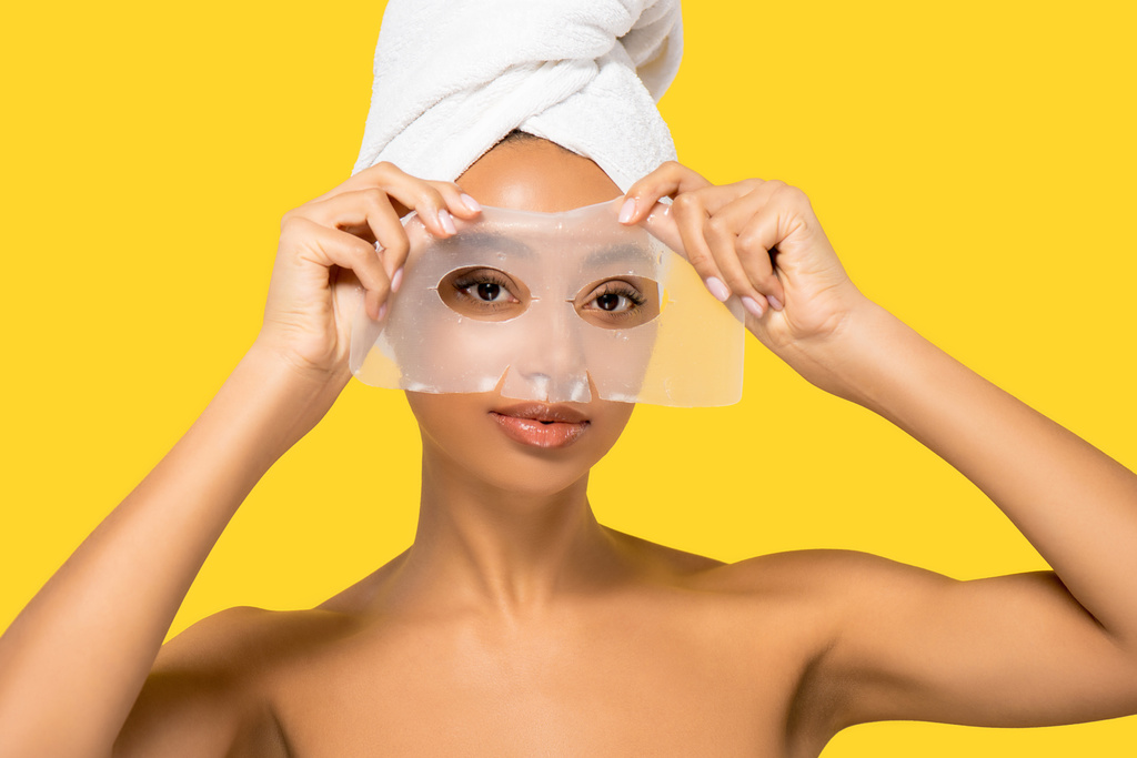 Afrikaans Amerikaans meisje met handdoek op het hoofd van toepassing hydrogel masker, geïsoleerd op geel - Foto, afbeelding