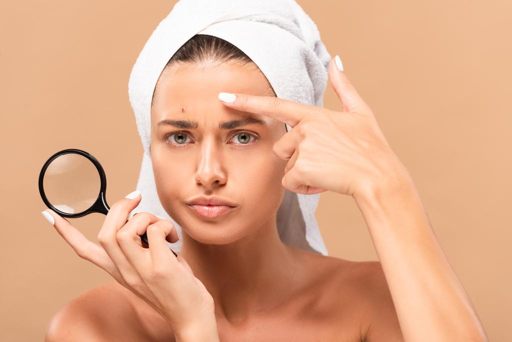 displeased girl pointing with finger at pimple on face and holding magnifier isolated on beige  - Photo, Image
