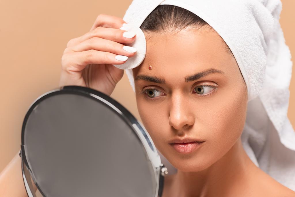 mladá žena drží bavlněné podložky při pohledu na zrcadlo izolované na béžové  - Fotografie, Obrázek