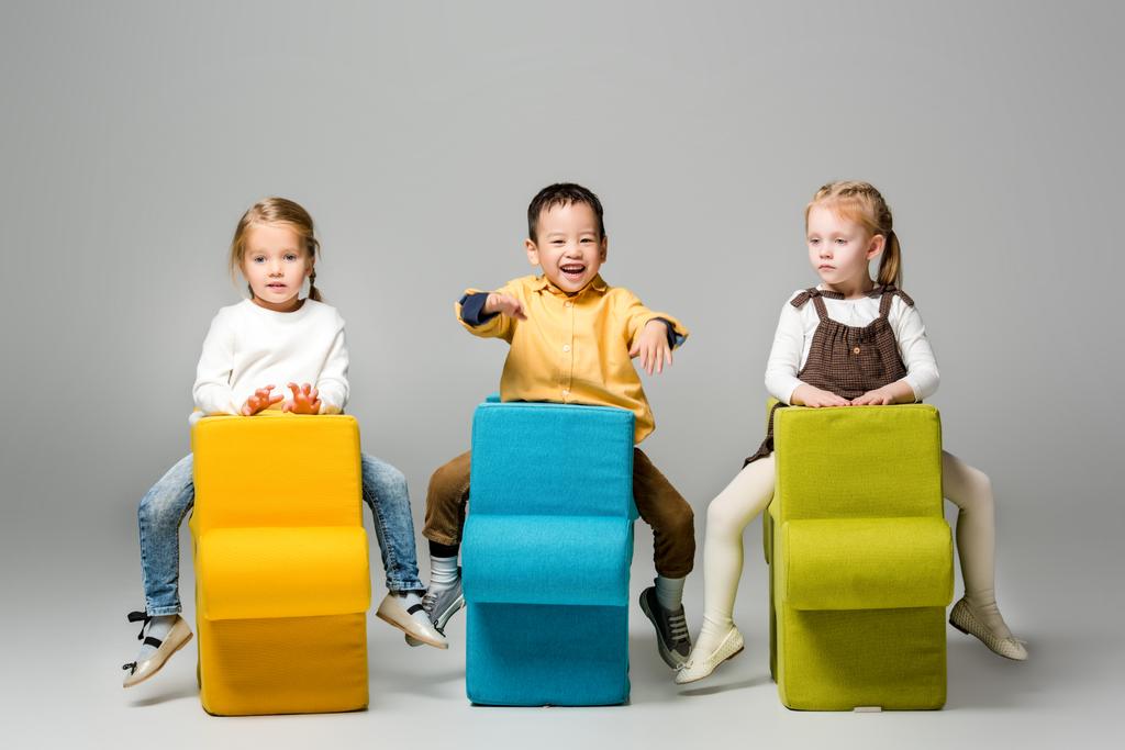 szczęśliwy wielokulturowe dzieci siedzi na krzesła puzzle, na szary - Zdjęcie, obraz