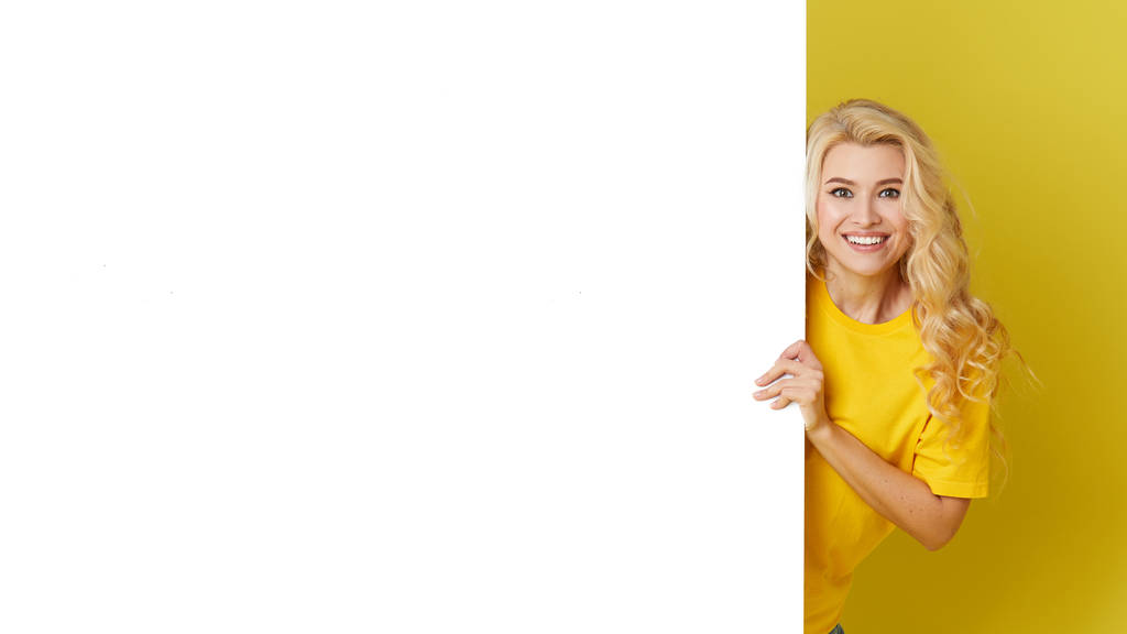 Joven mujer feliz se asoma por detrás de una pancarta blanca sobre un fondo amarillo. Apunte a un espacio vacío en un formulario, un espacio de copia para el texto. Disparo horizontal
 - Foto, imagen