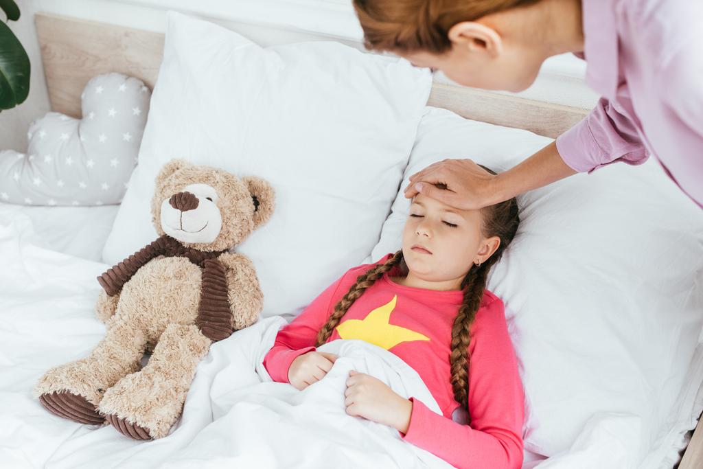 μητέρα αγγίζοντας το μέτωπο της άρρωστης νυσταγμένης κόρης στο κρεβάτι με αρκουδάκι - Φωτογραφία, εικόνα