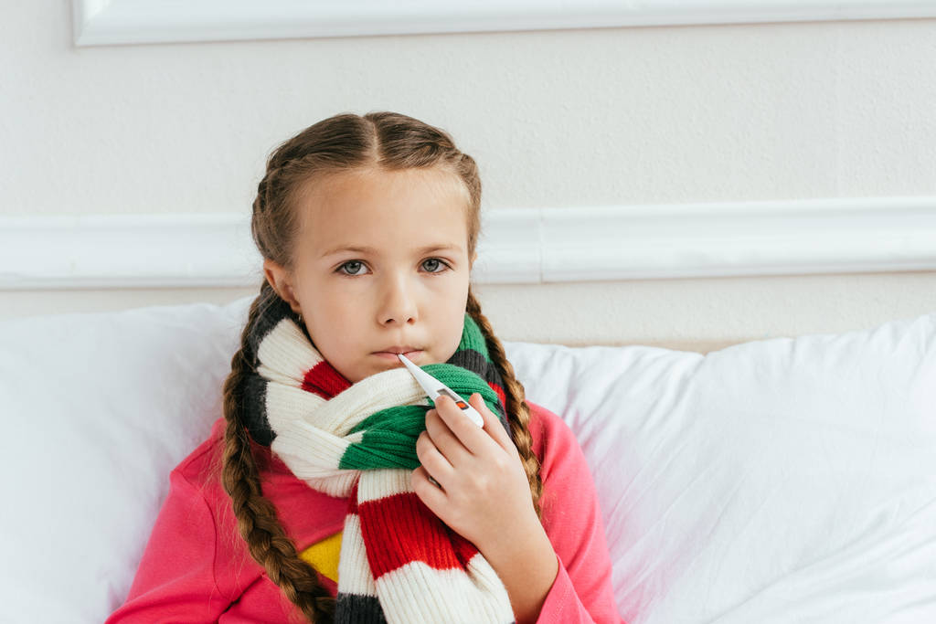 грустный больной ребенок в шарфе с лихорадкой и держа термометр на кровати
 - Фото, изображение