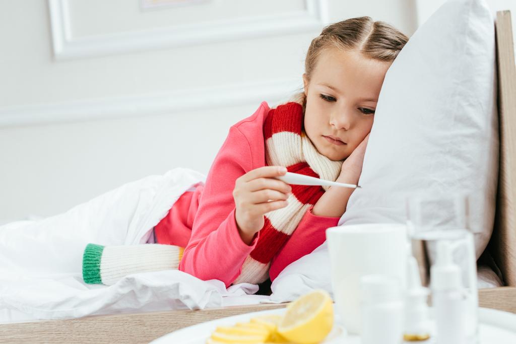 хвора засмучена дитина з гарячкою в шарфі, дивлячись на термометр, лежачи на ліжку з ліками поруч
 - Фото, зображення