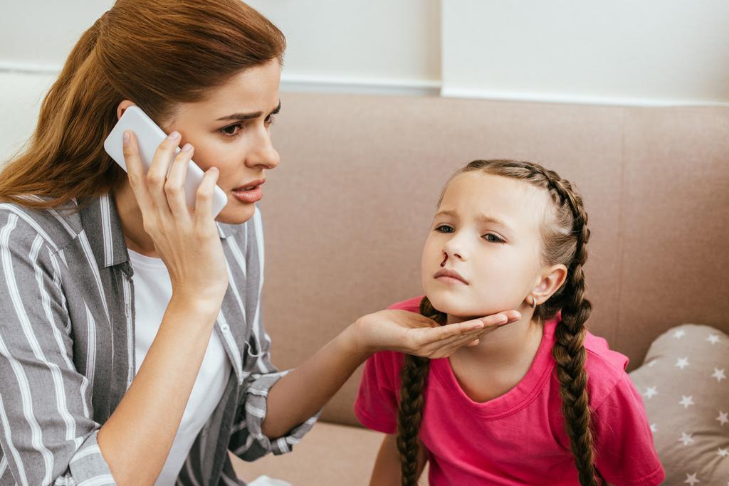 bezorgde moeder met online overleg op smartphone terwijl haar dochter neusbloedingen heeft - Foto, afbeelding