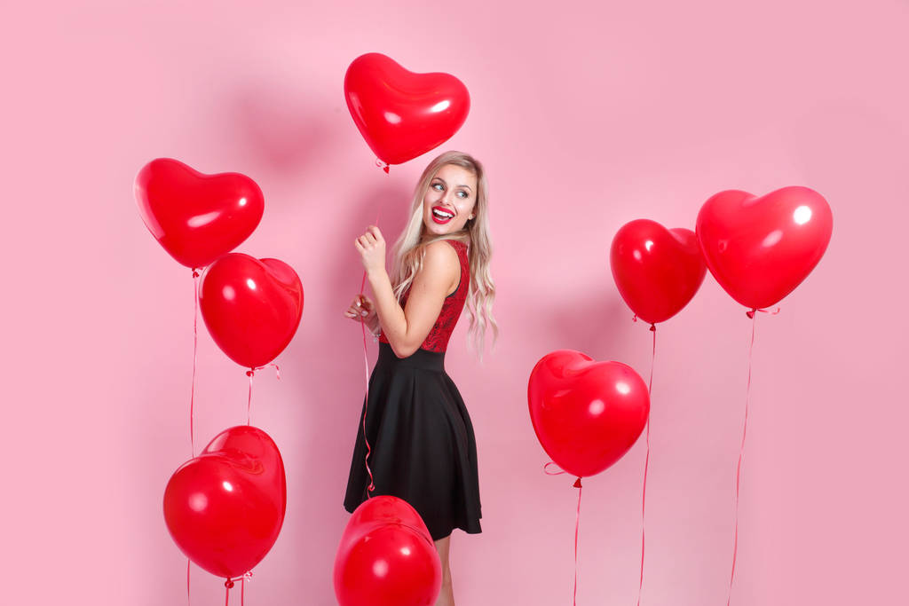 Mooie emotionele vrouw in elegante jurk met rode hart ballonnen op roze achtergrond - Foto, afbeelding