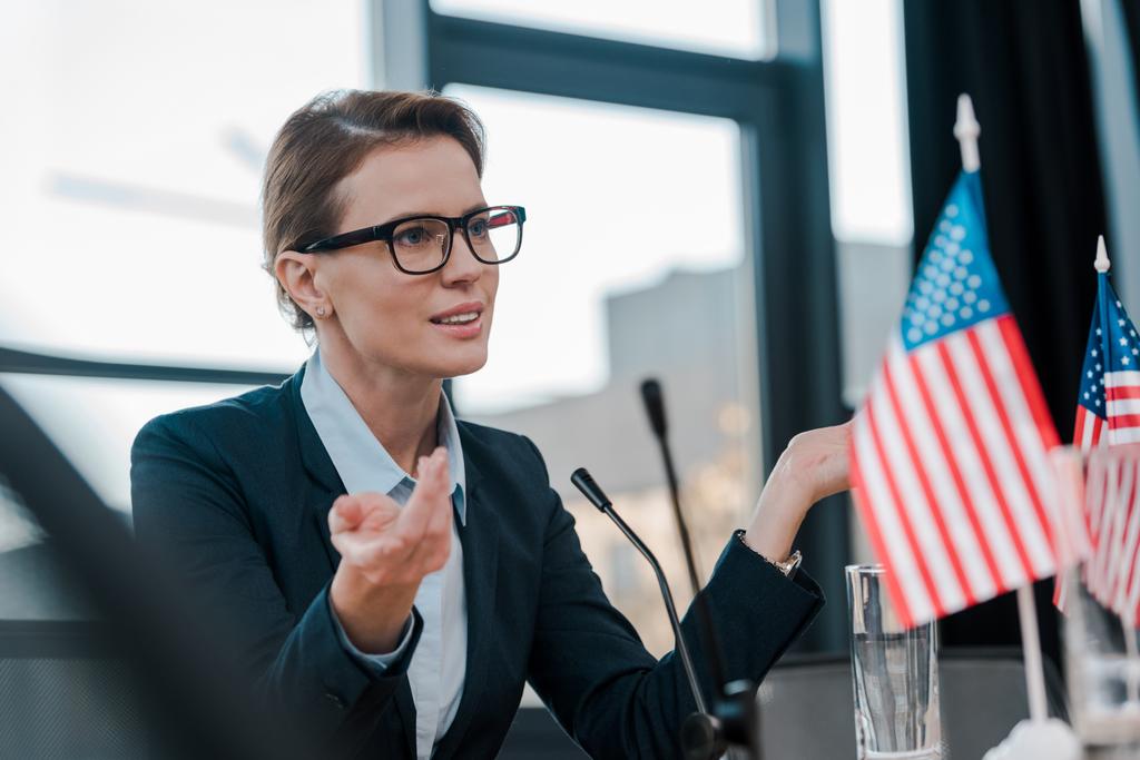 gyönyörű diplomata szemüveges gesztikulál, miközben beszél közelében mikrofon és amerikai zászlók  - Fotó, kép