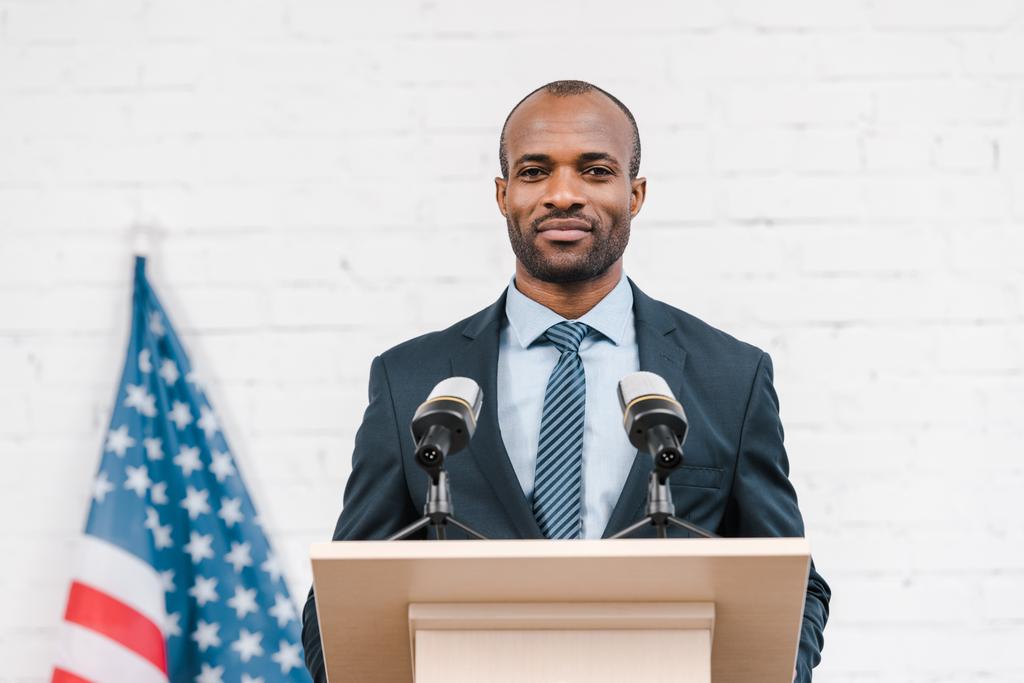 onnellinen afrikkalainen amerikkalainen puhuja seisoo lähellä mikrofonit ja Amerikan lippu
  - Valokuva, kuva
