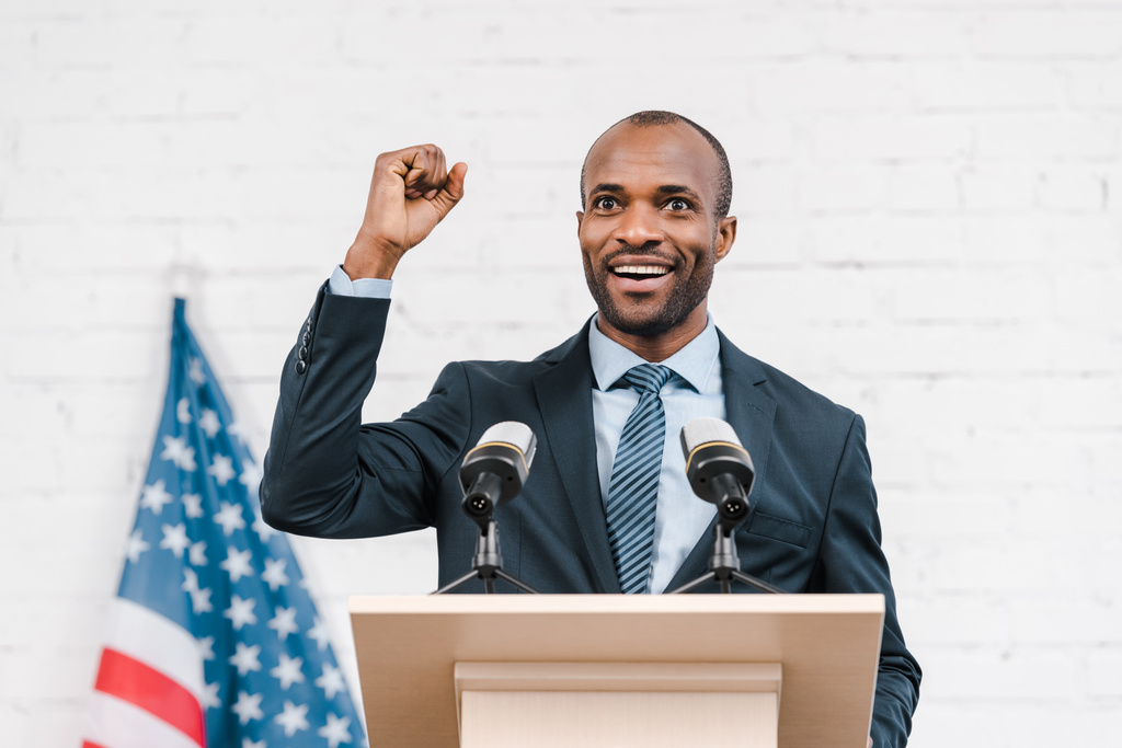 Χαρούμενος αφρικάνικος αμερικανός ομιλητής με σφιχτή γροθιά χαμογελώντας κοντά σε μικρόφωνα και αμερικανική σημαία  - Φωτογραφία, εικόνα
