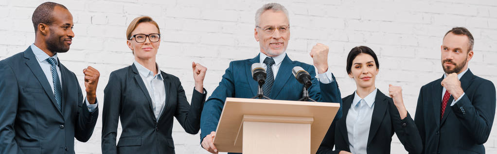 панорамный снимок счастливого оратора в очках и мультикультурных бизнесменов и бизнесвумен, показывающих сжатые кулаки
  - Фото, изображение