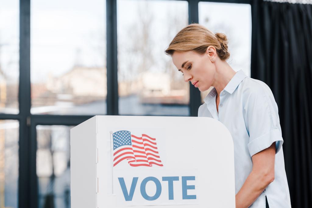 Seitenansicht einer attraktiven Frau, die in der Nähe eines Standes mit Wahlschriftzug und amerikanischer Flagge abstimmt  - Foto, Bild