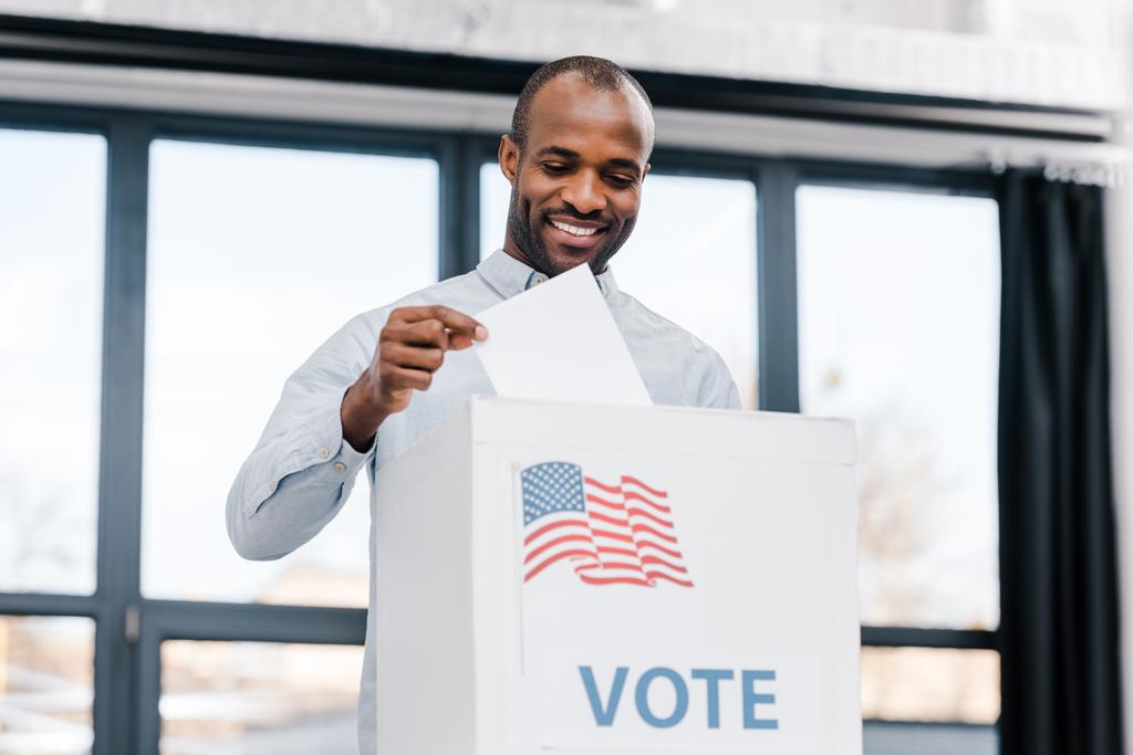  homem americano africano feliz votando e colocando cédula em caixa com bandeira da América
 - Foto, Imagem