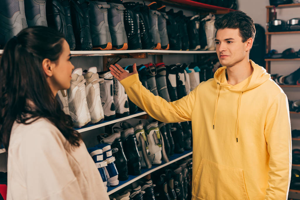 работник указывает рукой на лыжные ботинки и смотрит на клиента в ремонтной мастерской
  - Фото, изображение