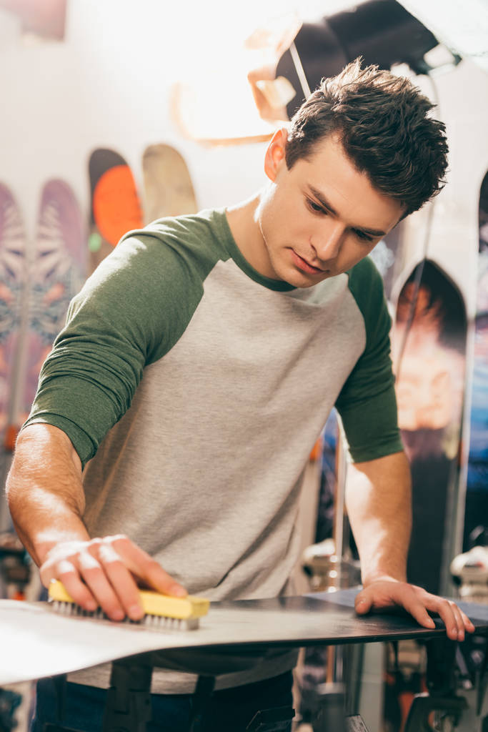 handsome worker using brush on snowboard in repair shop  - Photo, Image